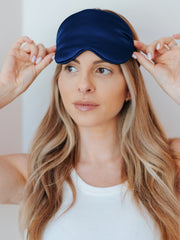Woman wearing a navy blue satin sleep mask against a plain background