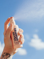 A bottle of hydrating face and body mist being held up to the bright sunny sky and sprayed. The hydrating mist spray can be seen in the warm summers air.